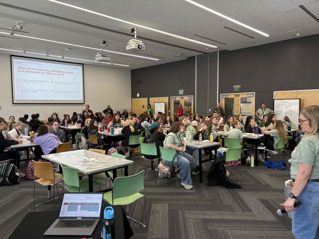 Students sit at tables in a large conference room while presenters lead a literacy competition session from the front.