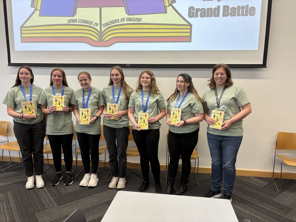 Students and a teacher wearing medals and matching shirts hold books after participating in the Iowa Grand Battle reading competition.