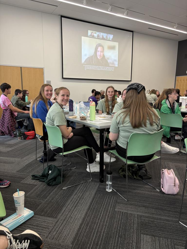 Students sit at tables during a literacy conference while a speaker appears on a large screen at the front of the room.