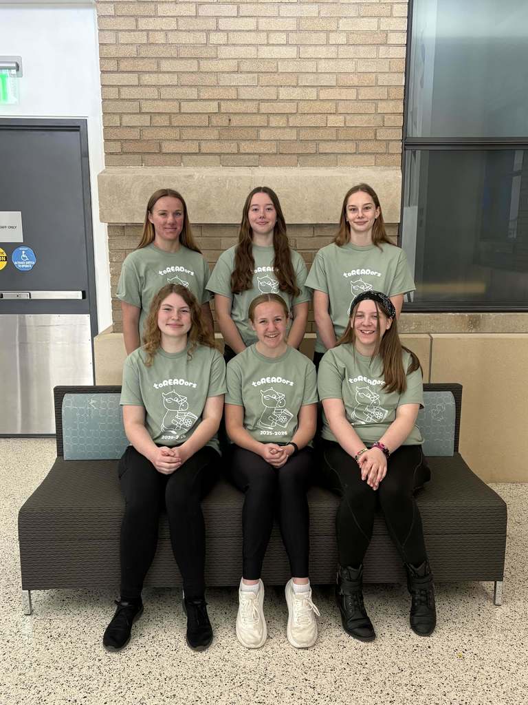 Six students wearing matching “toREADors” shirts pose together indoors against a brick wall during a reading competition event.