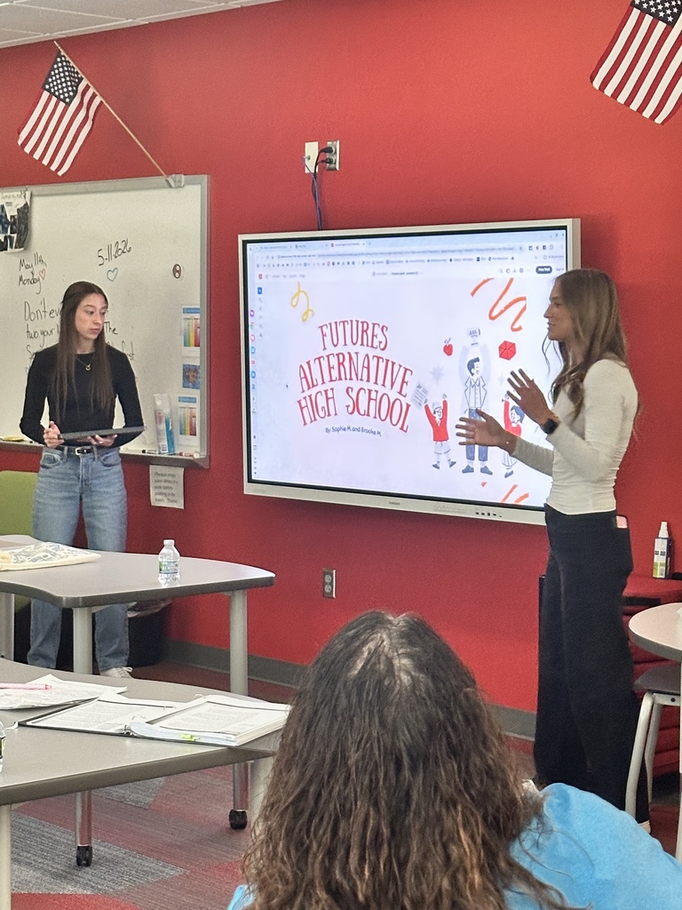 Two students present “Futures Alternative High School” on a classroom screen while classmates listen.