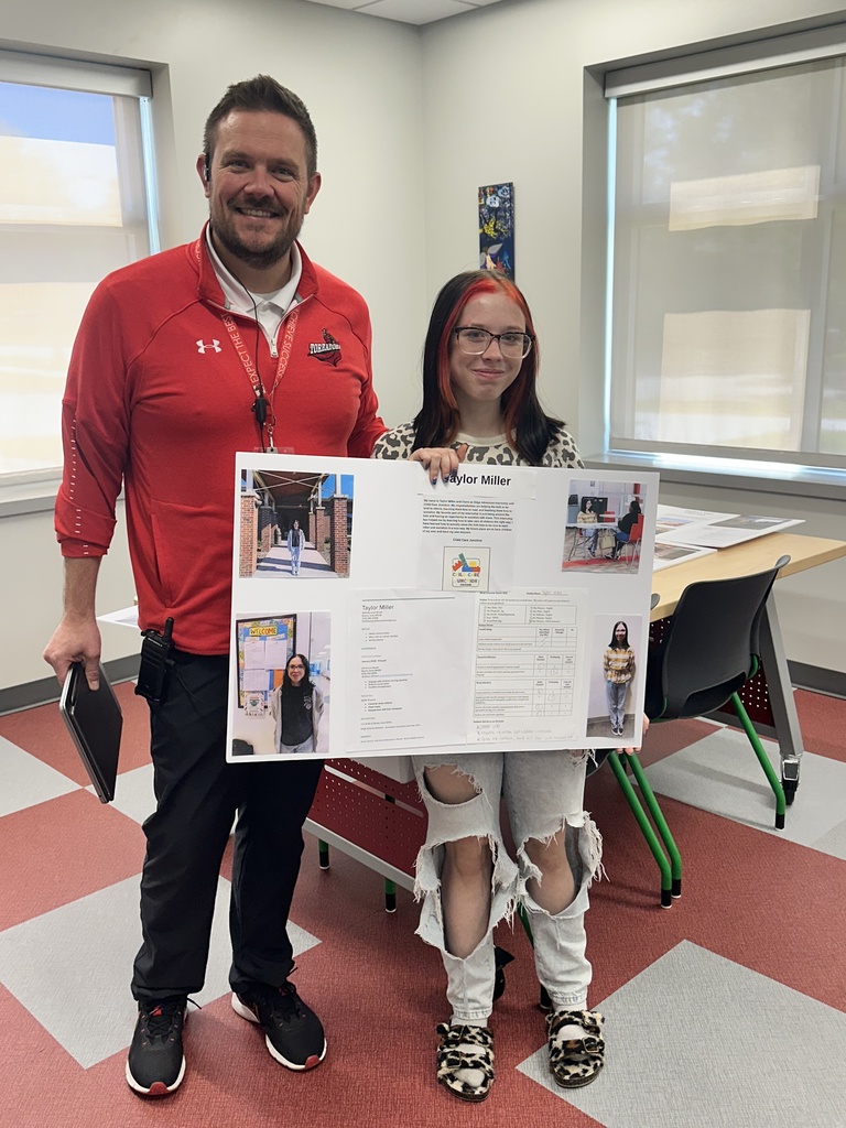 Two people standing in a classroom holding a large presentation board with text and photos.