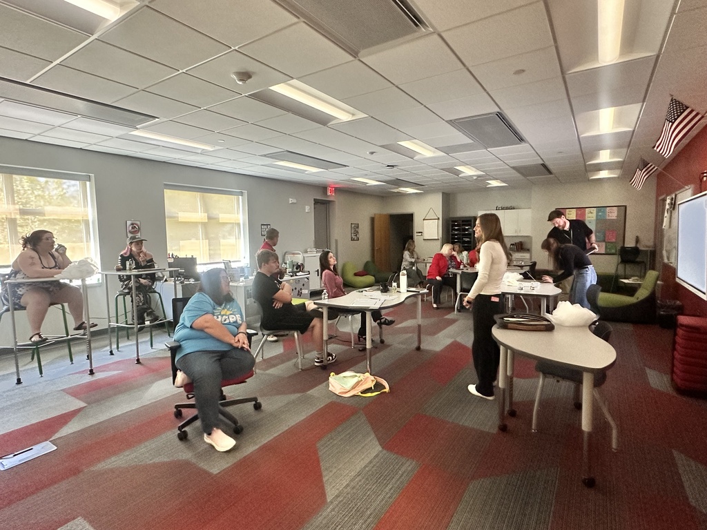 Wide view of a classroom discussion with students and staff seated around tables in a bright learning space.