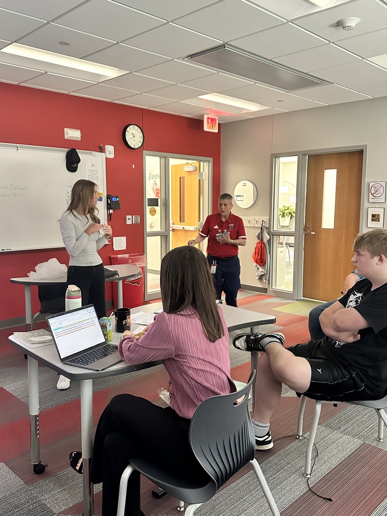 Students sit at tables in a classroom while two presenters speak near a whiteboard and digital display.