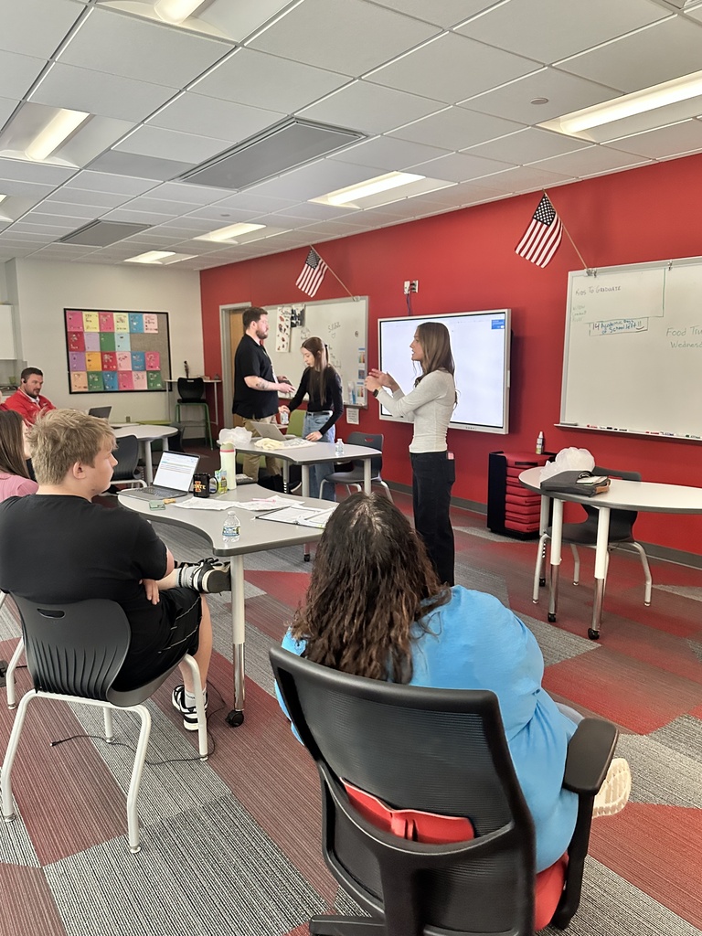 Presenter speaks to seated students in a classroom with red accent walls, whiteboards, and American flags.