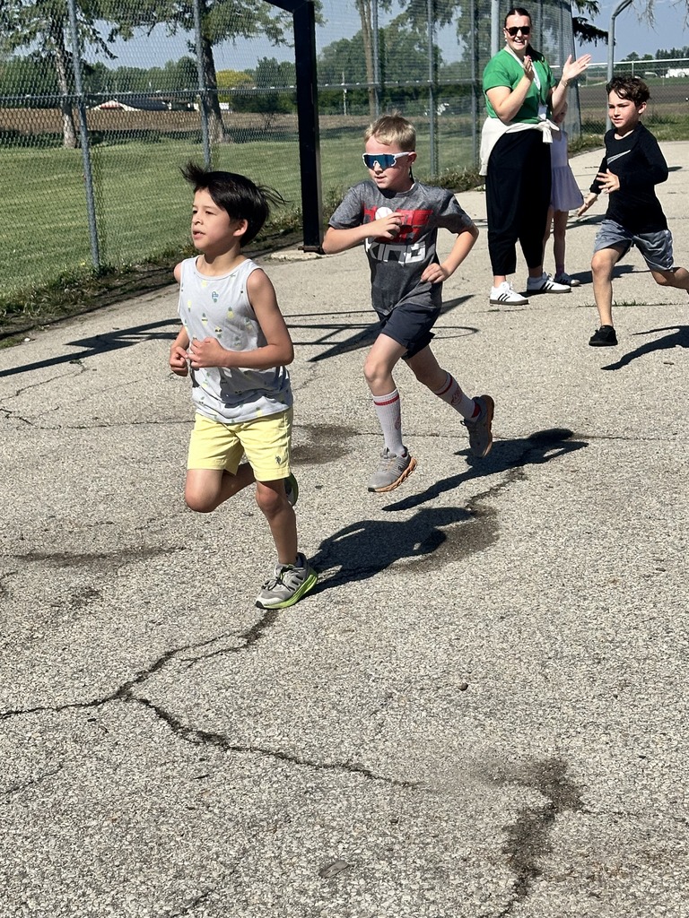 Young students run on a paved school path during an outdoor race while a teacher cheers from the sidelines.