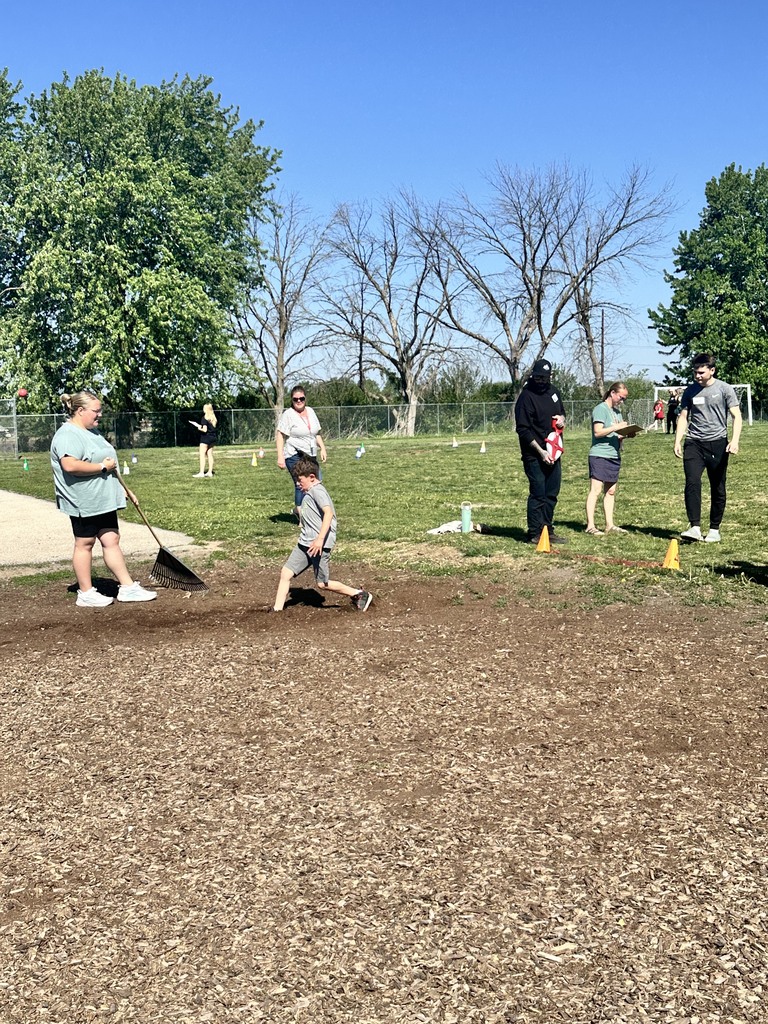 A young boy runs on a dirt field while adults stand nearby, some holding rakes, under a clear blue sky with trees in the background.