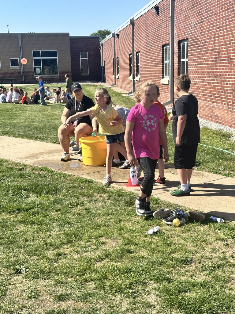 Elementary students play a water balloon game outside school buildings while classmates and staff watch nearby.