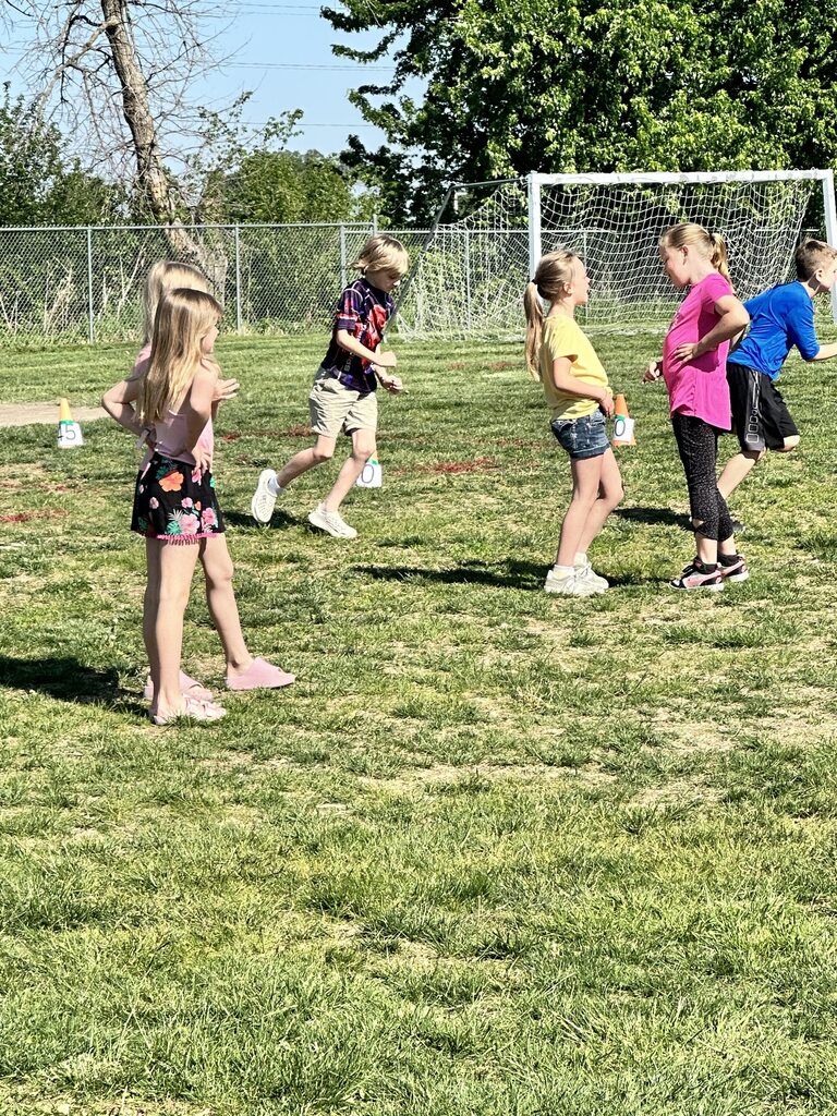 Elementary students stand and move across a grassy field during an outdoor school activity near soccer goals.
