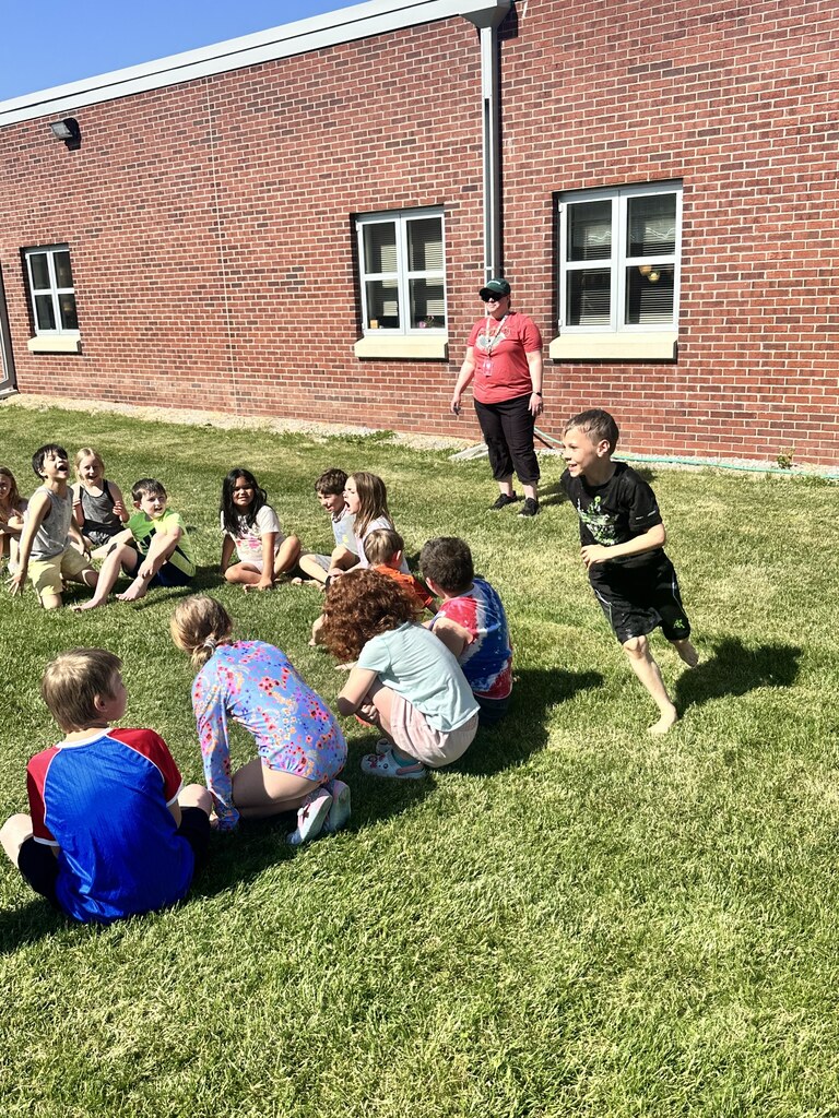 Children sit and play on the grass outside a school building while a teacher supervises nearby.