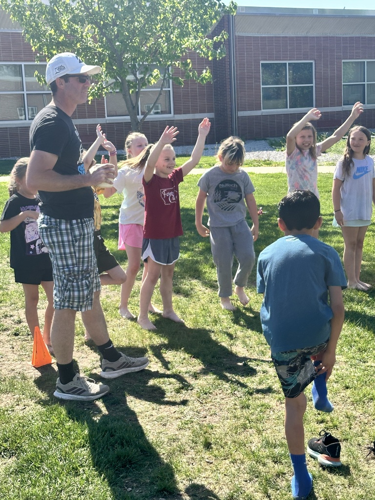 Children raise their hands during an outdoor school game led by an adult on a grassy field.