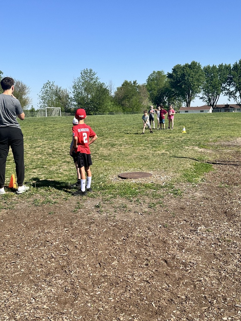 Children playing on a sunny field with an adult observing nearby under a clear blue sky.