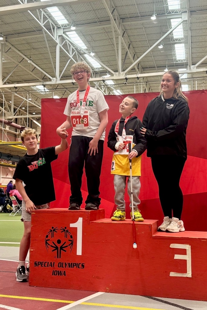 Four athletes stand on a Special Olympics Iowa podium inside a gym, celebrating their achievements with medals and smiles.
