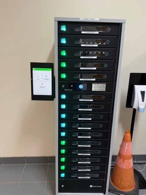 A tall storage unit with multiple compartments and a screen is mounted on a wall. Adjacent is a traffic cone.