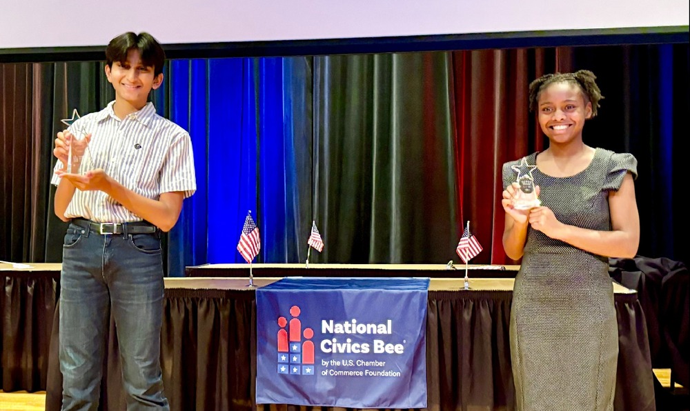 Photo of Om Ganapathy and Kamilyah Woodrow standing on a stage, holding trophies in front of a National Civics Bee banner. 
