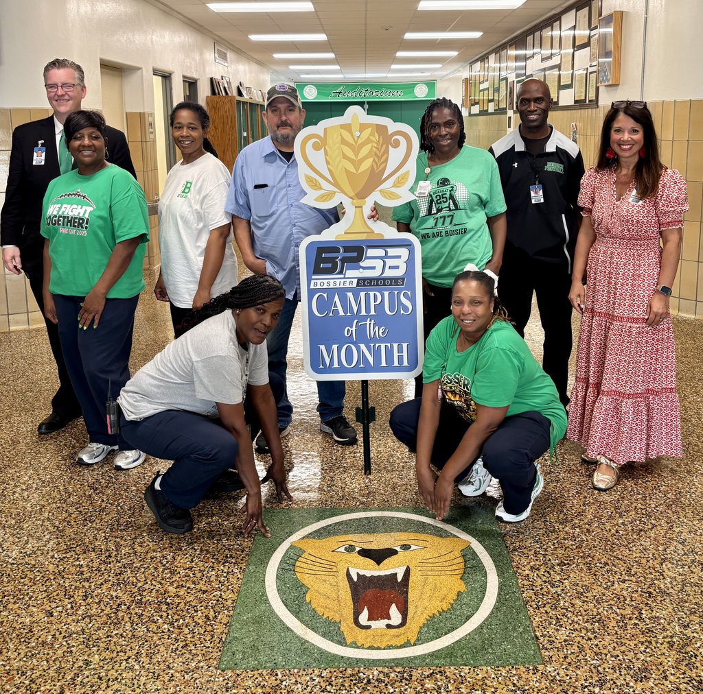 Group photo inside a Bossier High School hallway celebrating the Campus of the Month award. Nine school staff members and district administrators gather around a large sign featuring a gold trophy graphic and the text "BPSB Bossier Schools Campus of the Month." Two staff members kneel in front next to a floor decal of the green and gold Bearkat mascot, while Assistant Superintendent Andrea Spinney stands on the far right in a patterned red dress.