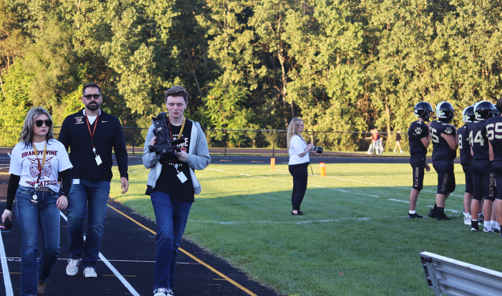 two kids with a microphone and camera walking on the track with their teacher at football game