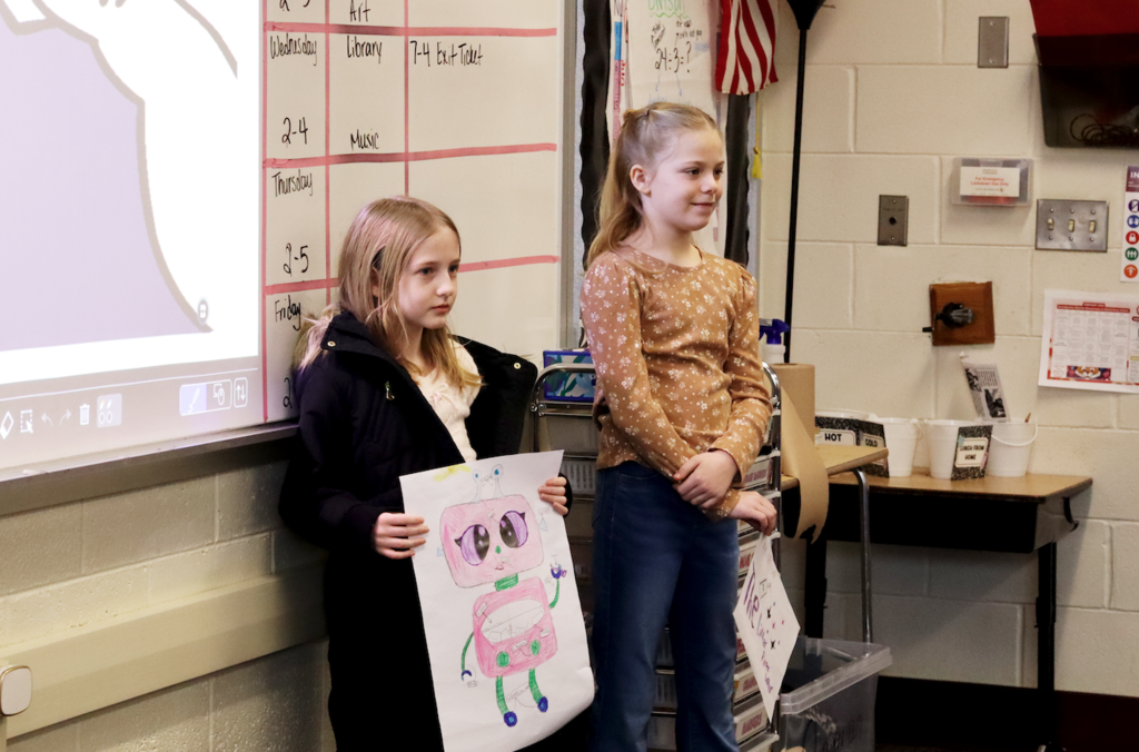 two girls presenting, one girl holding picture of robot