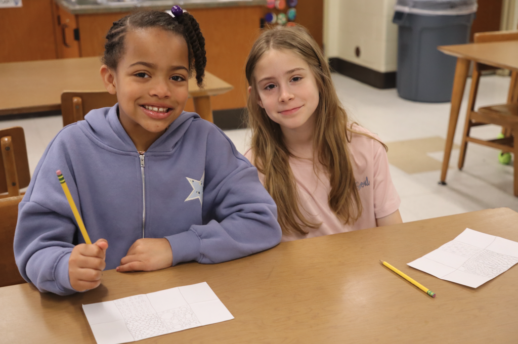 two young girls in art class smiling at camera with papers and pencils in front of them