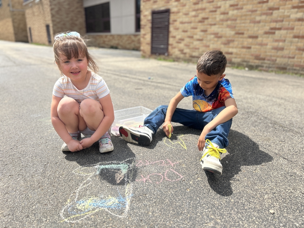 little girl smiling at camera with little boy sitting on the pavement, playing with sidewalk chalk