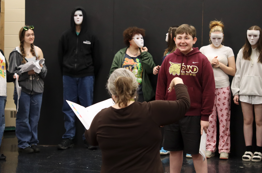 young man standing on stage smiling, while teacher directs