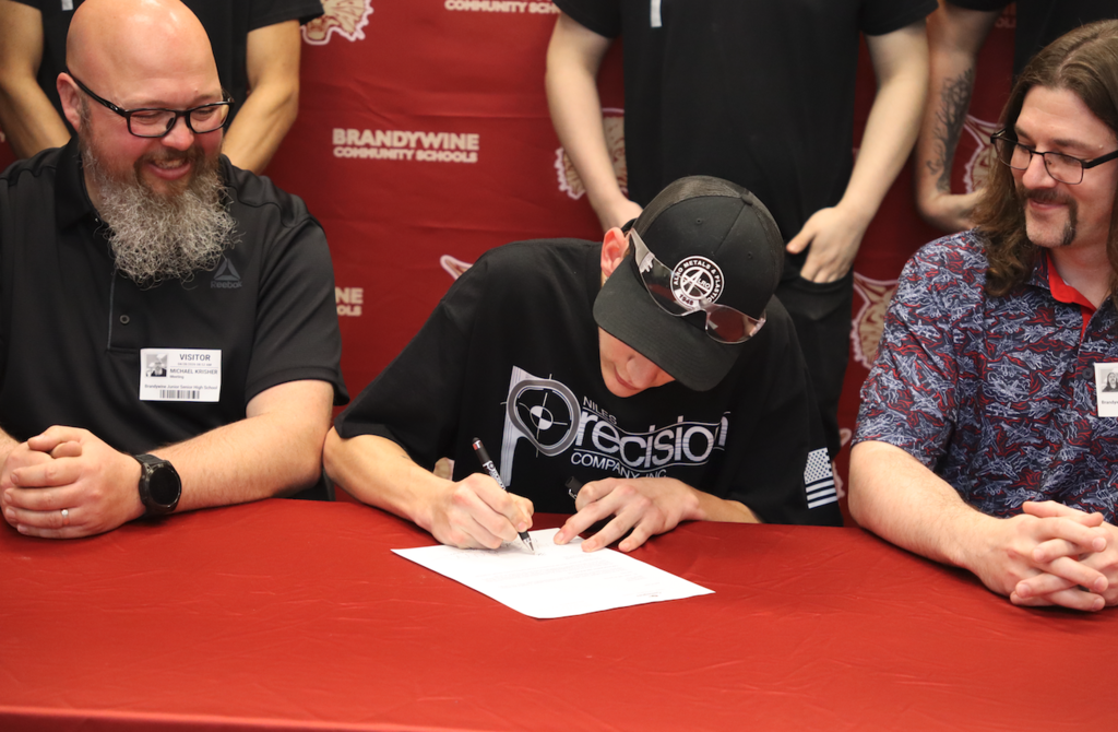 students sitting with two men at a table signing for apprenticeship