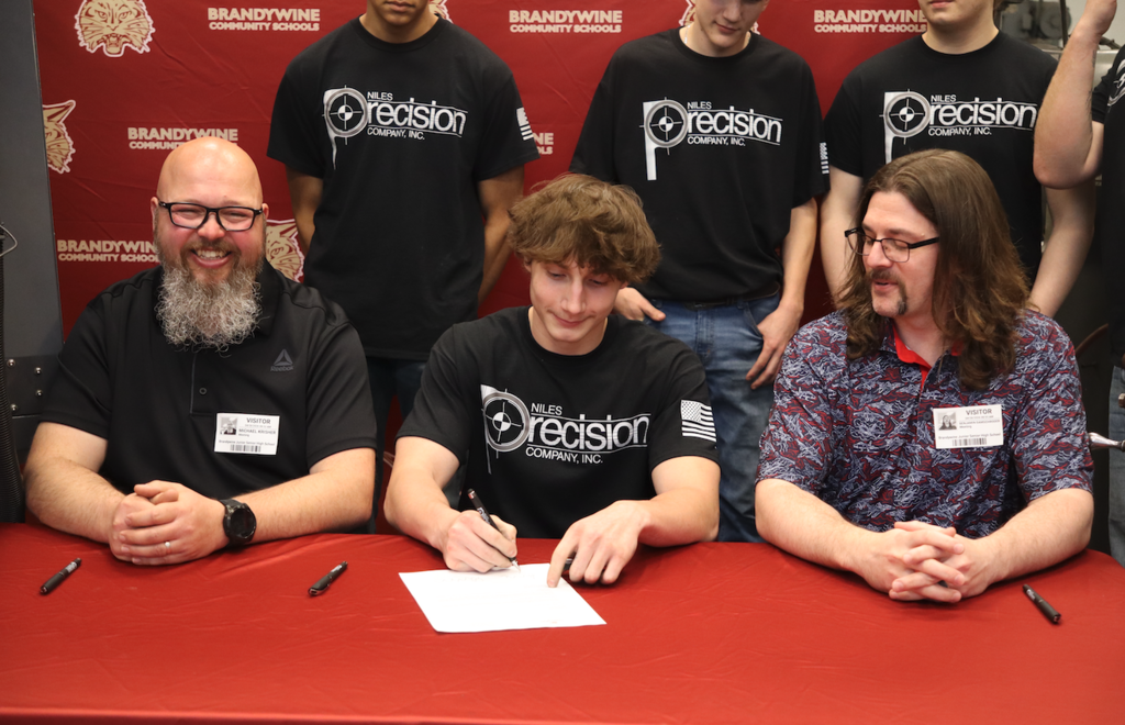 students sitting with two men at a table signing for apprenticeship
