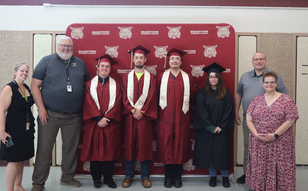 four graduates standing in front of backdrop in caps and gowns with teachers