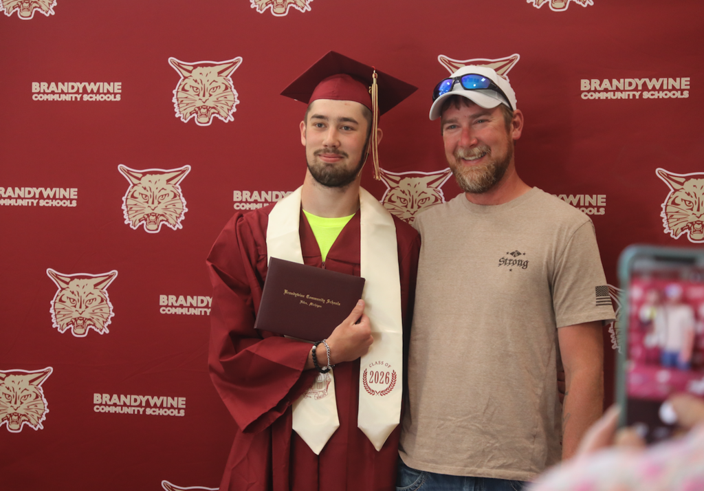 graduate smiling in front of bobcat backdrop with another male, holding his diploma