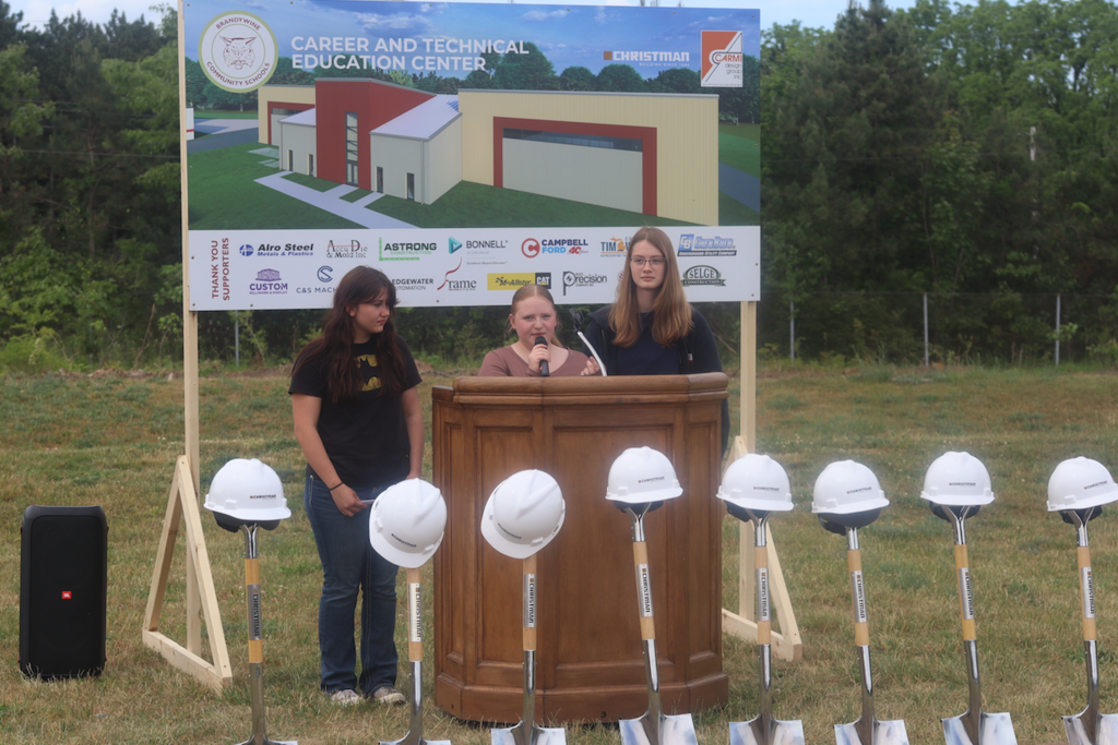 students speaking at groundbreaking with hard hats and shovels in front of them