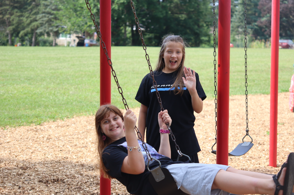 one girl swinging, with another girl waving at the camera