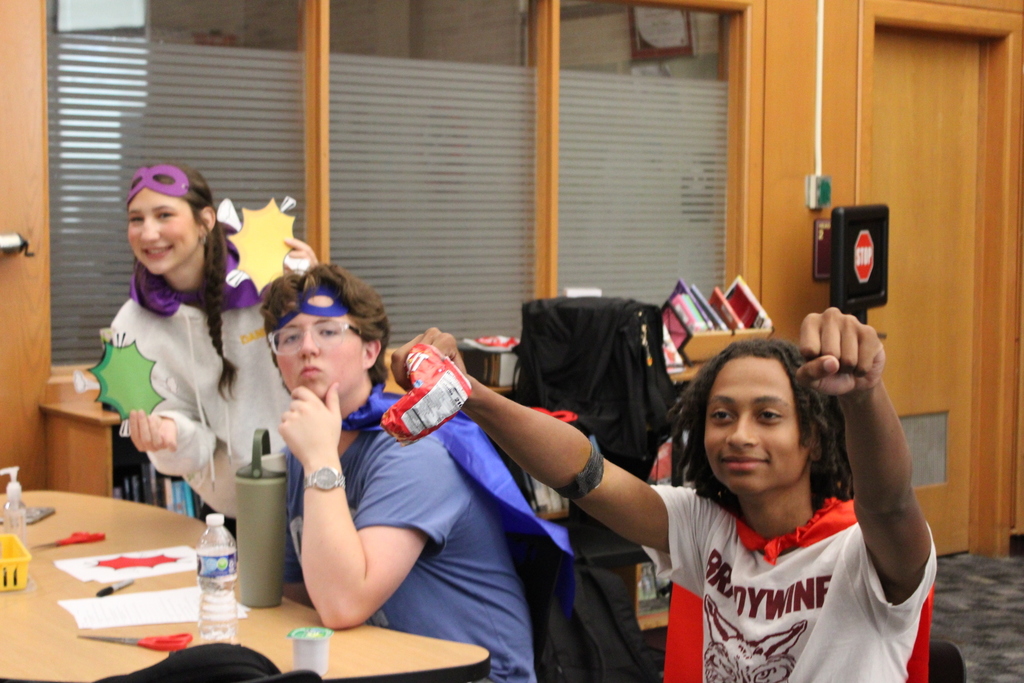 students in superhero costumes sitting at table, with arms up
