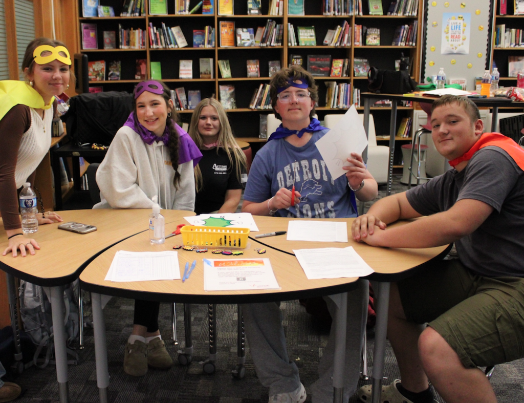 group of five students sitting at table at blood drive wearing superhero outfits