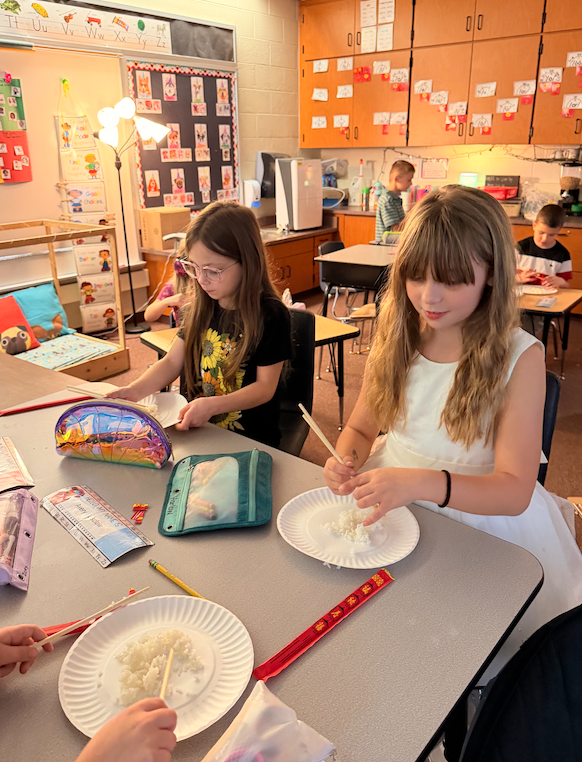 two girls sitting at desk trying to eat rice with chopsticks