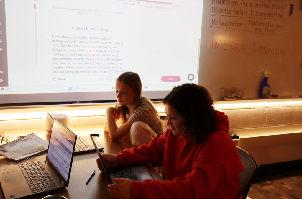 two girls sitting in front of laptops with book on screens