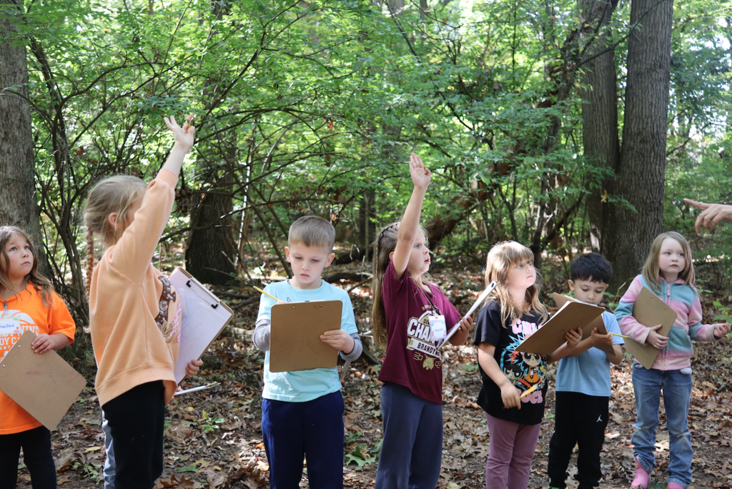 kids standing outside in the woods with hands up and clipboards