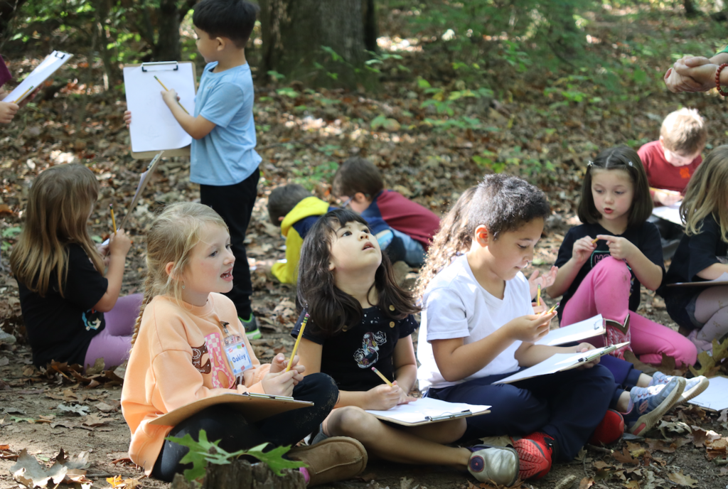 class of kindergarteners sitting on the ground with clipboards