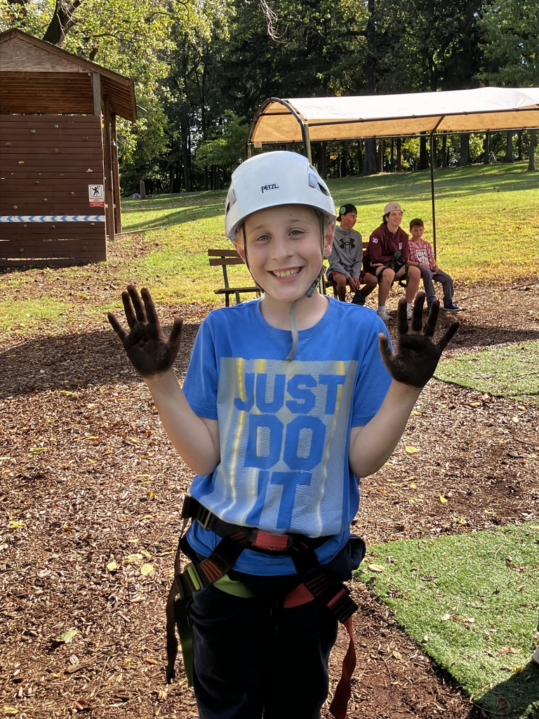 student stands in harness next to climbing activity with muddy hands