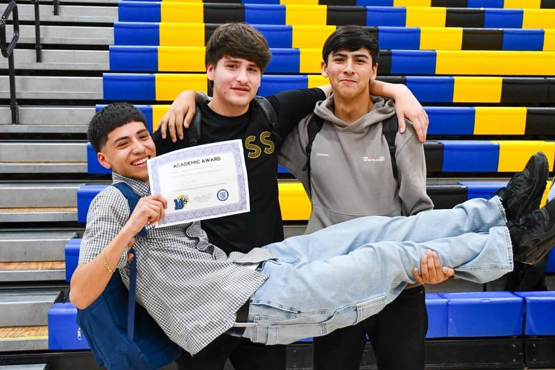 Two students hold up another student while he holds certificate at award assembly.