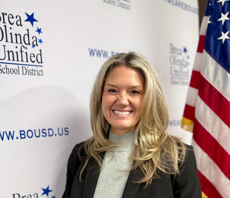 Trustee Natalie Johnson stands next to an American Flag and in front of a BOUSD backdrop. She is wearing a gray turtleneck and black blazer
