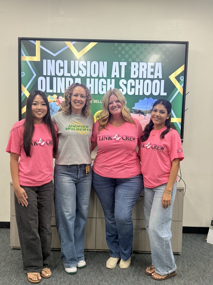 2 students in pink shirts stand with 2 educators from BOHS. Behind them there is a TV monitor with a green and gold graphic that reads Inclusion at Brea Olinda High School