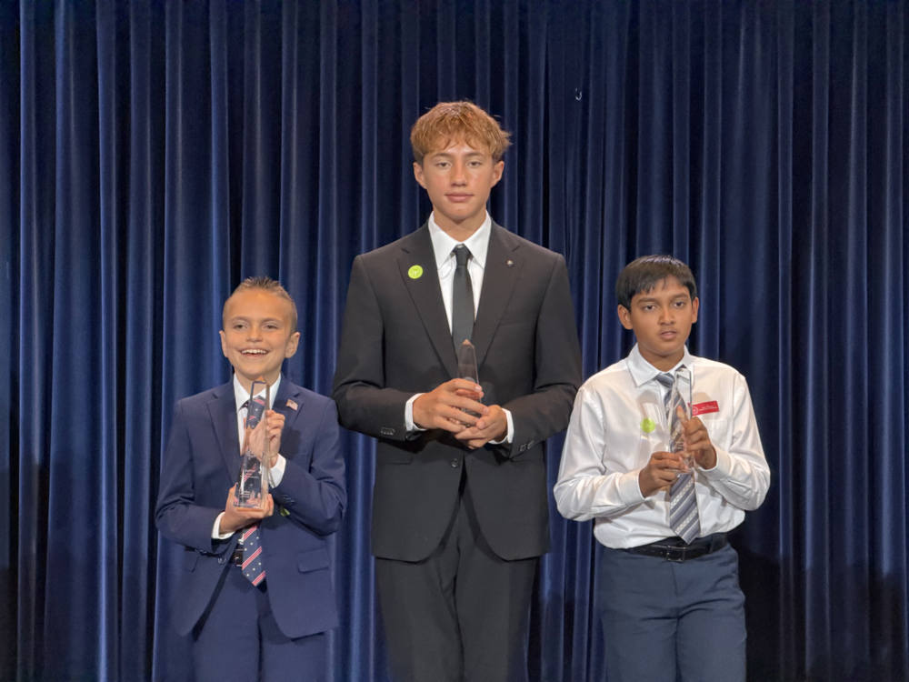 three students stand in front of a dark blue curtain holding awards