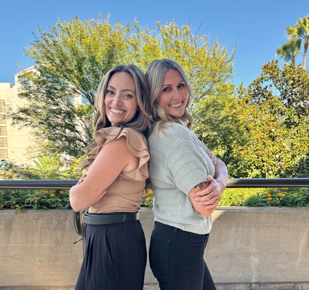 two women stand back to back with trees behind them