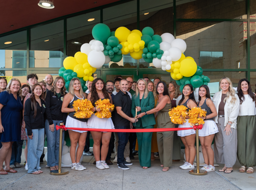 a group of people stand behind a red ribbon while one person in the middle holds large scissors. Balloon arch above them in green, gold and white
