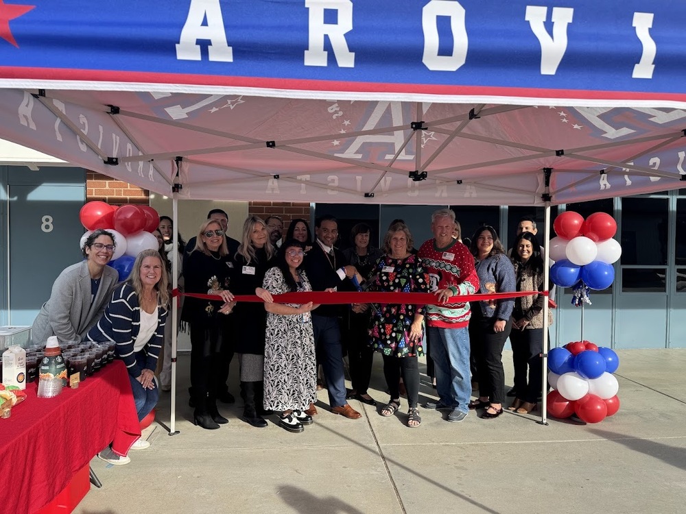 a group photo of people standing under an Arovista tent and behind a red ribbon. Someone in the middle is holding a pair of large scissors and ready to cut the ribbon