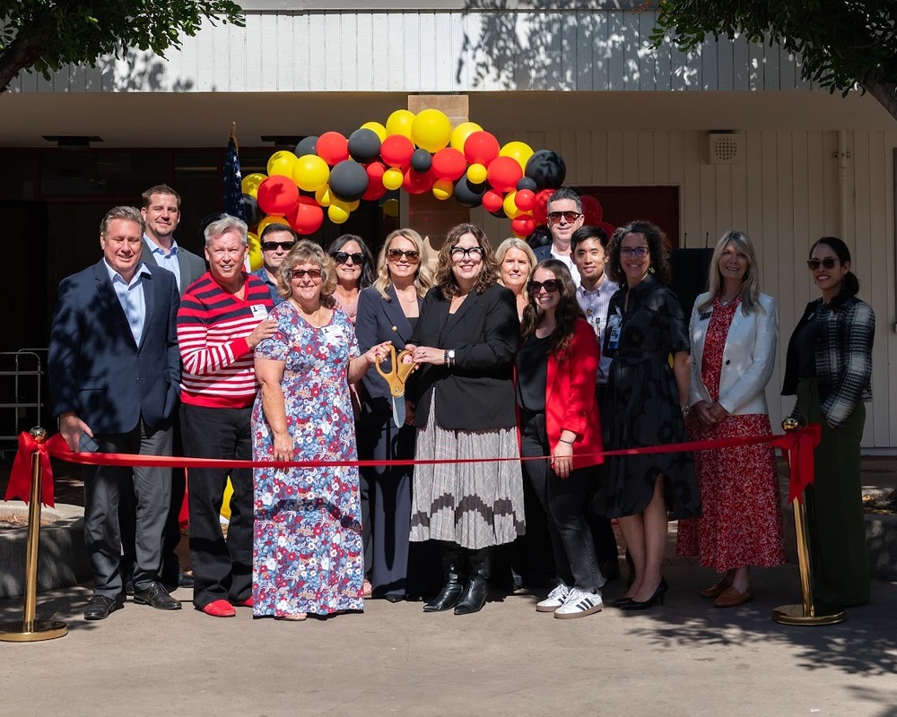 a group of people stand under a balloon arch and in front of a red ribbon. and they are holding large scissors
