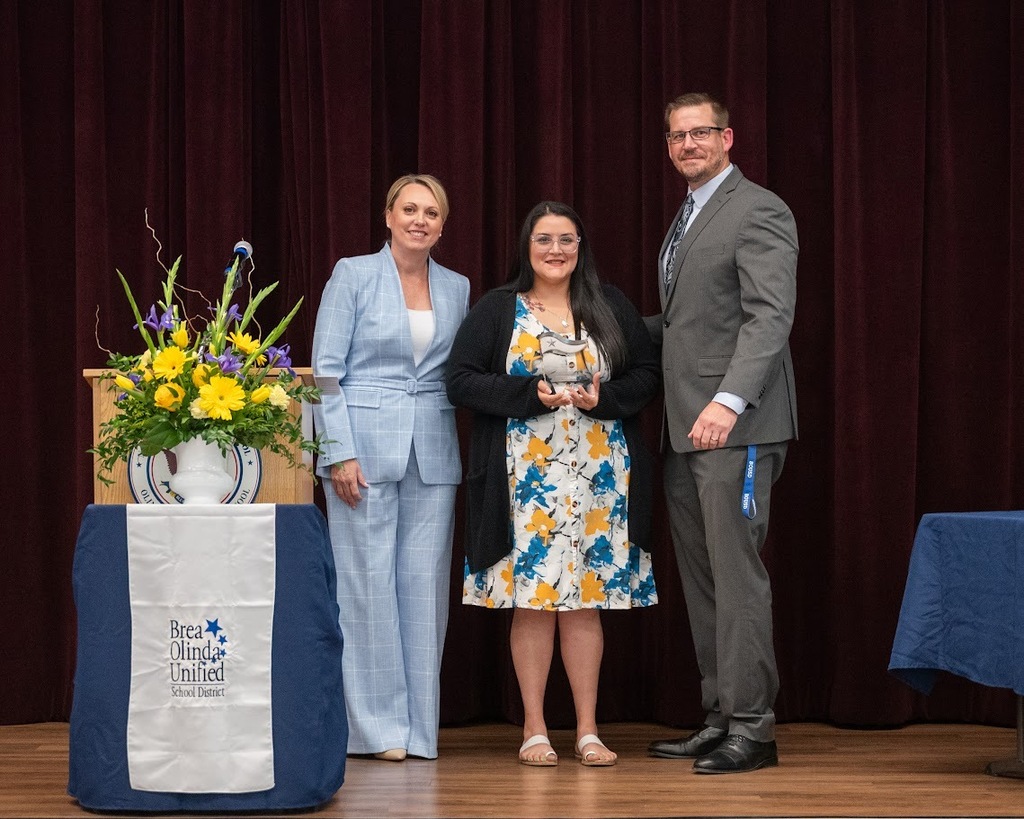 brinda leon stands with ms. molina and assistant superintendent josh porter. Ms Molina holds her award. They are all standing next to a podium with a white bousd banner covering the podium and flowers on top