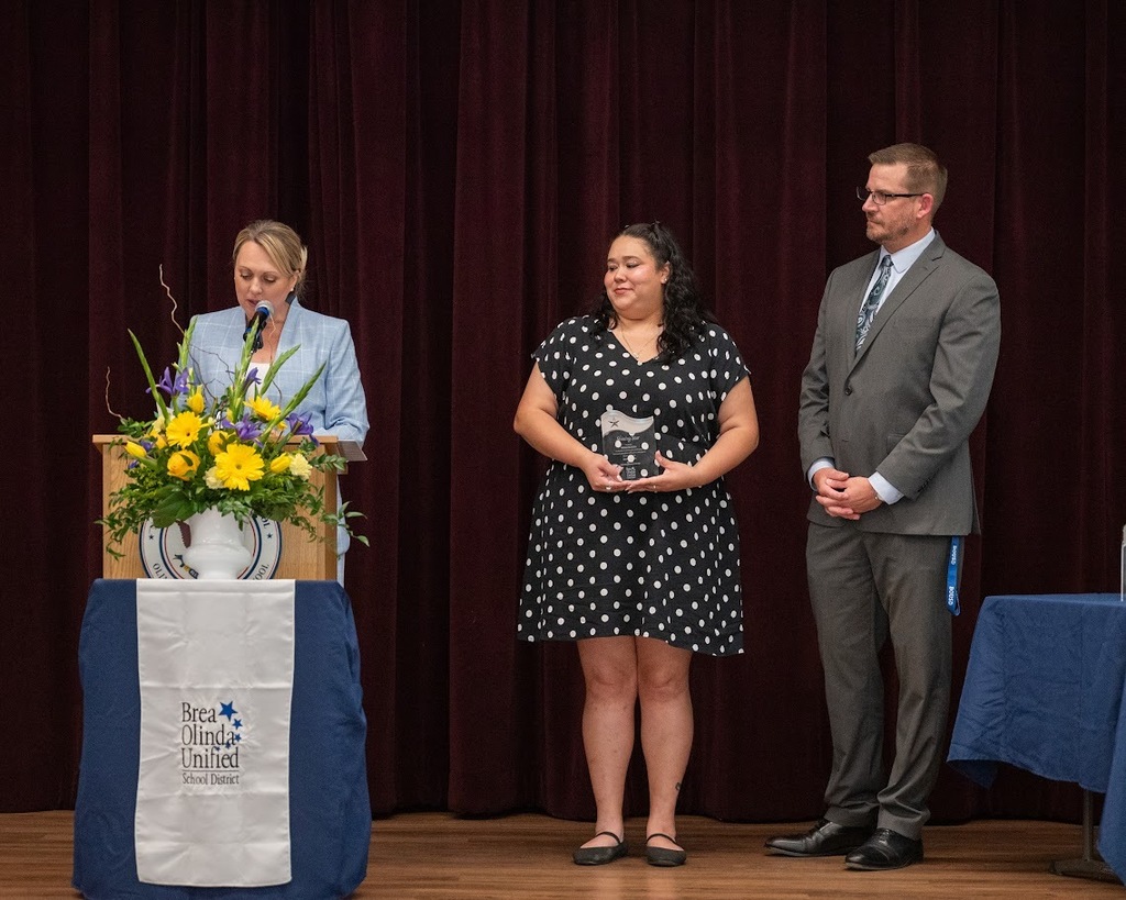 Ingrid palacios stands on stage with her award as superintendent leon speaks at podium. 