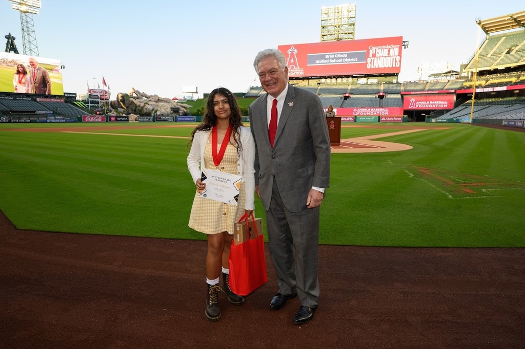 Camila stands with a gentleman dressed in a dark gray suit on the baseball field of Angel Stadium
