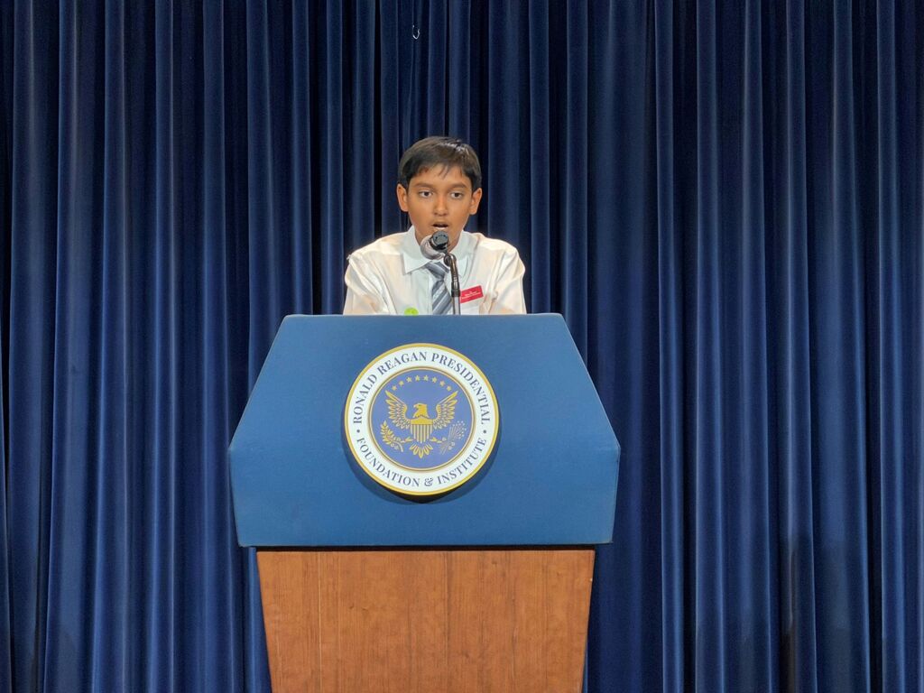 Aryan speaks into a microphone at the podium with the presidential seal and blue curtain behind